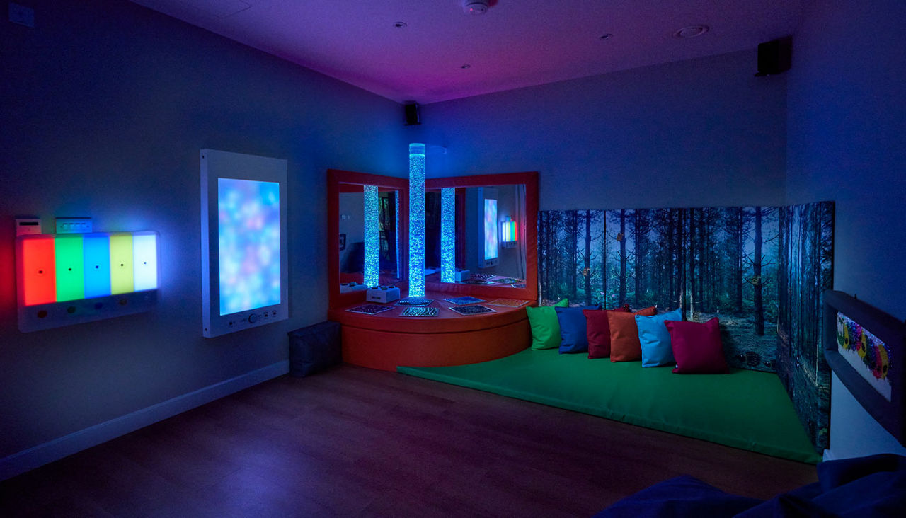 Two illuminated bubble tubes glow blue, reflecting in mirrors, while LED panels pulse multicolors. Context: a dim sensory room with a green mat, colorful cushions, and a forest-wall mural.