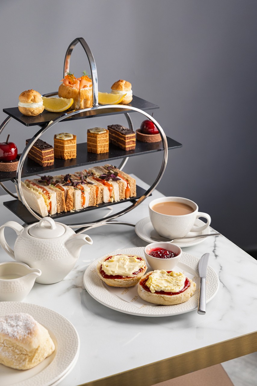 afternoon tea cake stand with cakes, and sandwiches next to a tea pot, a cup of tea and a plate with scones