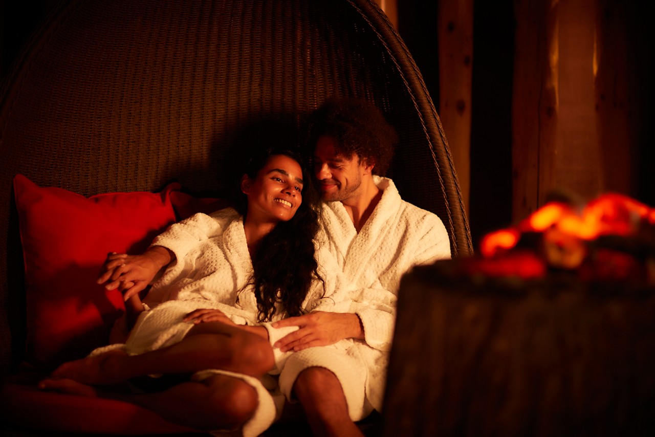 Man and woman snuggled up in a large cocoon chair by a glowing fire.
