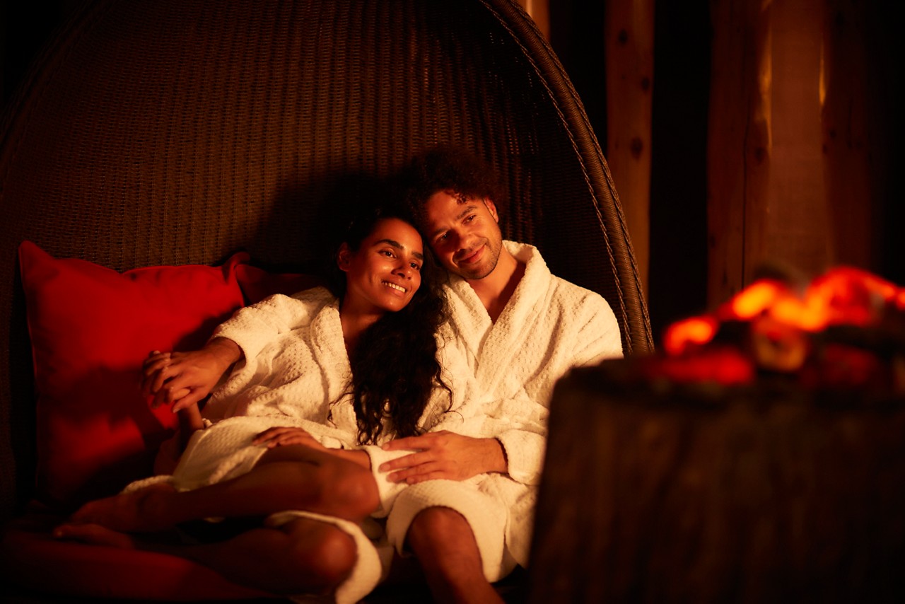 Man and woman snuggled up in a large cocoon chair by a glowing fire.