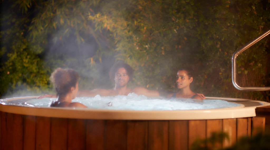 Three people relax and chat, sitting in a round, bubbling hot tub. Steam rises around them. Context: outdoor nighttime garden, leafy backdrop and metal handrail beside the wooden-sided tub.