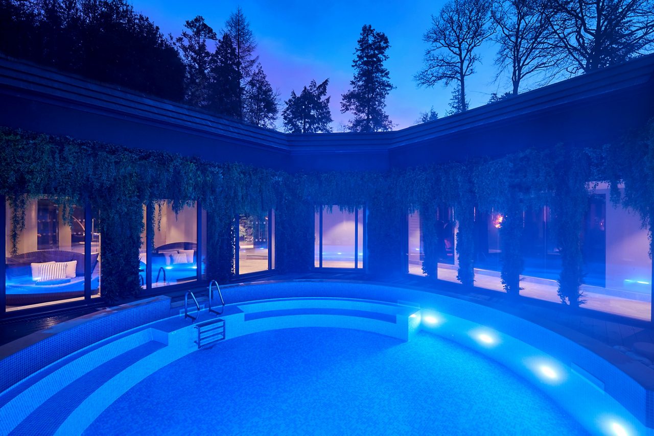 Curved swimming pool glows blue, lights washing steps and benches. Surrounding building with large windows and hanging greenery encloses a courtyard, while tall trees silhouette against twilight sky.