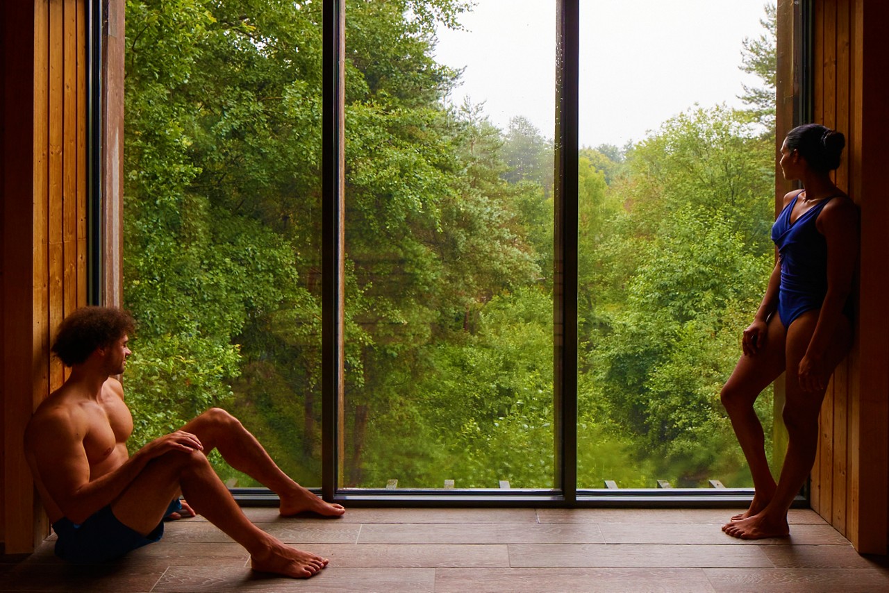 Man and a woman looking out at the forest from inside the Treetop Sauna.