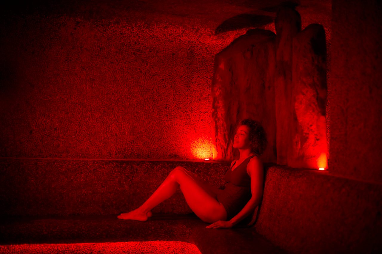 Person in a swimsuit reclines on a stone bench, eyes closed, illuminated by red lamps, inside a cave-like steam room with rough walls and a recessed light on the floor.