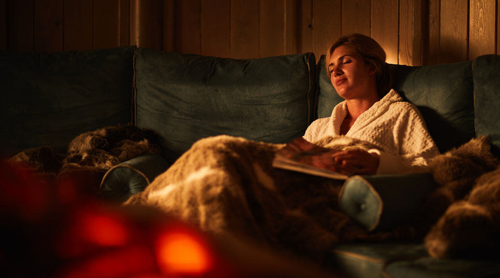 Person reclines on a teal sofa, eyes closed, holding a magazine under a furry blanket; warm firelight flickers in foreground within a wood-paneled, dim, cozy room.