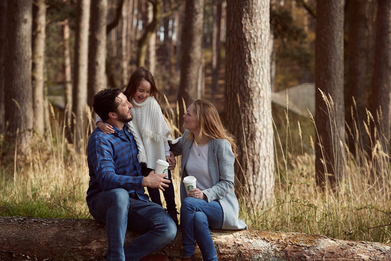 A father, daughter and mother sat on a log in the forest with starbucks coffee cups
