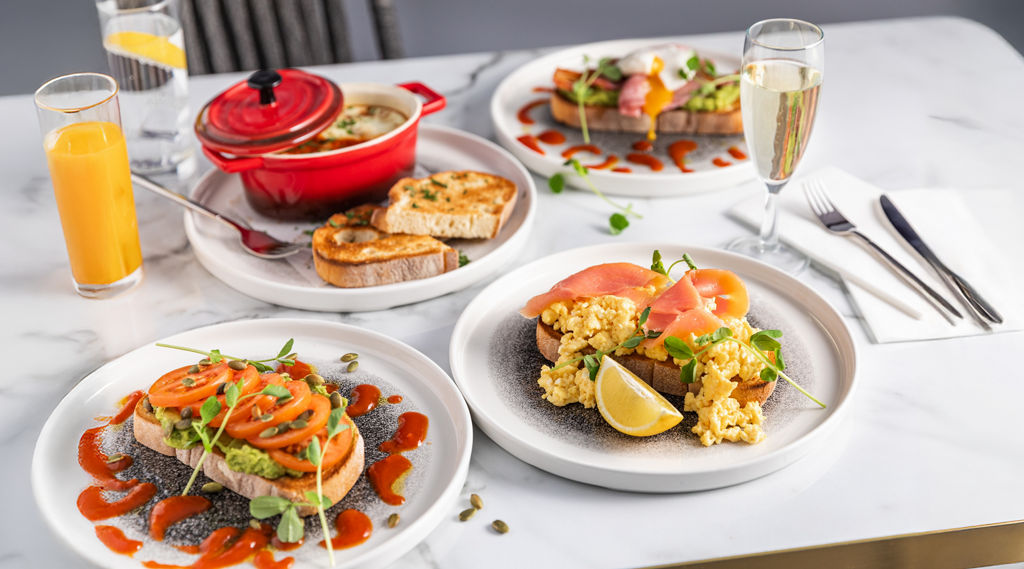 Brunch plates show toast topped with avocado, tomatoes, or scrambled eggs and smoked salmon, drizzled with sauce. Nearby: pot with bread, orange juice, water, champagne, and cutlery on a marble table.