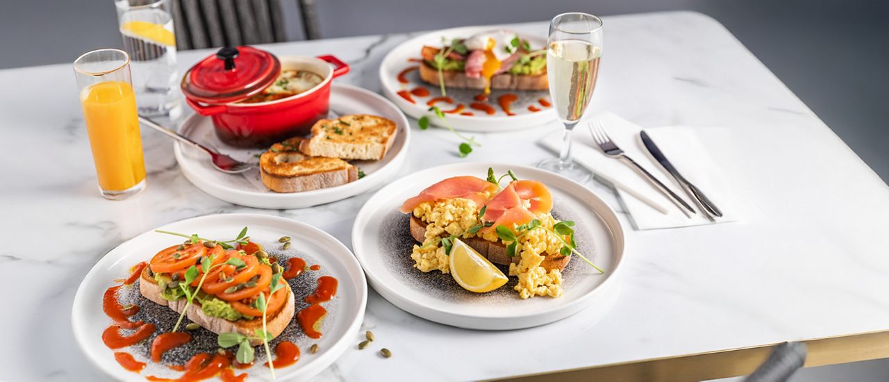 Brunch plates show toast topped with avocado, tomatoes, or scrambled eggs and smoked salmon, drizzled with sauce. Nearby: pot with bread, orange juice, water, champagne, and cutlery on a marble table.