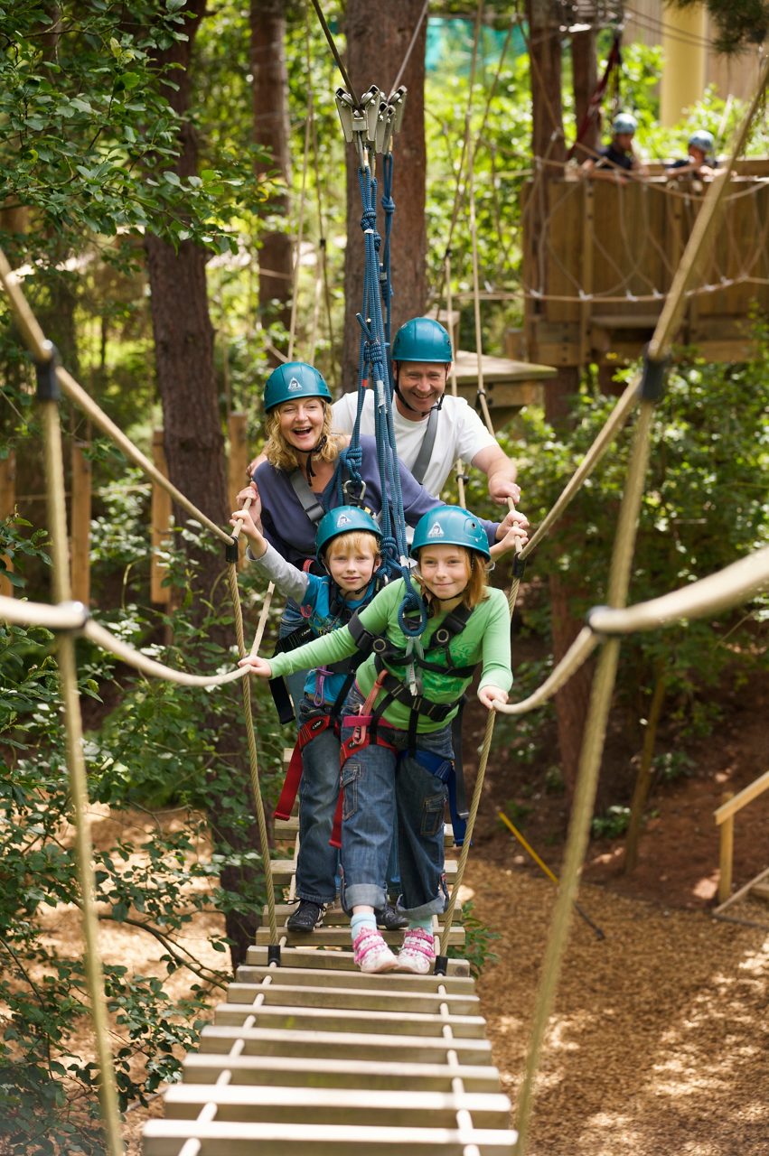 Two helmeted riders glide on parallel ziplines, legs outstretched and arms spread, suspended in harnesses, against a backdrop of tall green trees and overcast sky in an outdoor adventure park.
