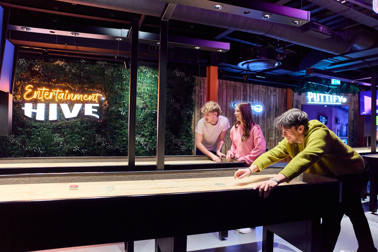 Three people play shuffleboard, sliding a puck down the table, in a neon-lit entertainment venue with greenery wall and signs. Text: "Entertainment HIVE", "PUTTIFY", "EXIT".
