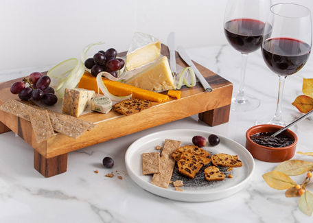 A wooden platter with different cheeses and a glass of red and white wine next to it