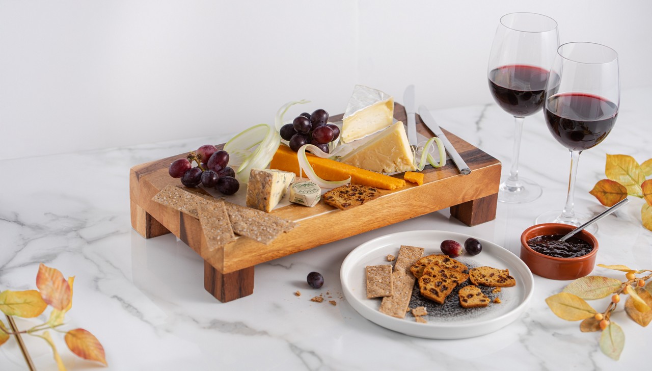 A wooden platter with different cheeses and a glass of red and white wine next to it