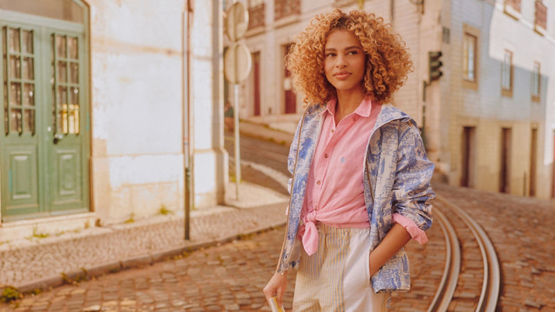A lady wearing a pink tshirt and patterned blue jacket stood in front of some buildings on a cobbled street. 