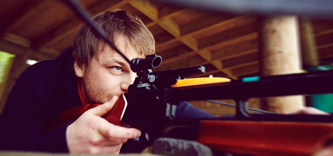 Scoped rifle aims forward; person sights through scope, cheek on stock, hands steady on grip. Wooden shelter background suggests a shooting range. Visible text: SAFE — FIRE.