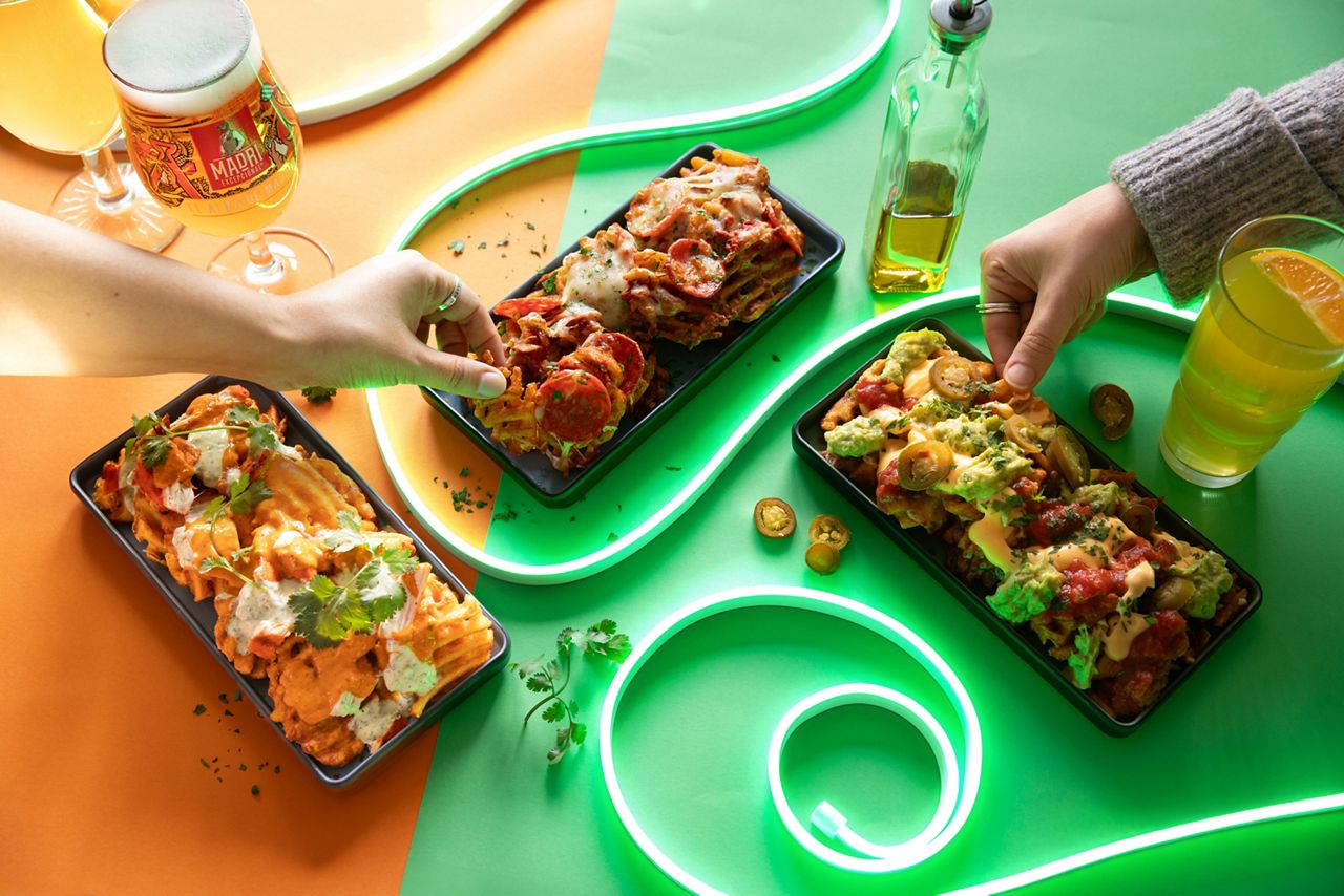 There are 3 plates of different flavour loaded waffle fries including pepperoni,  nacho and chicken tikka. There is a glass of Madri beer and a soft drink and they all sit on a brightly coloured green and orange background with led lighting. There are two different hands picking up some of the fries.