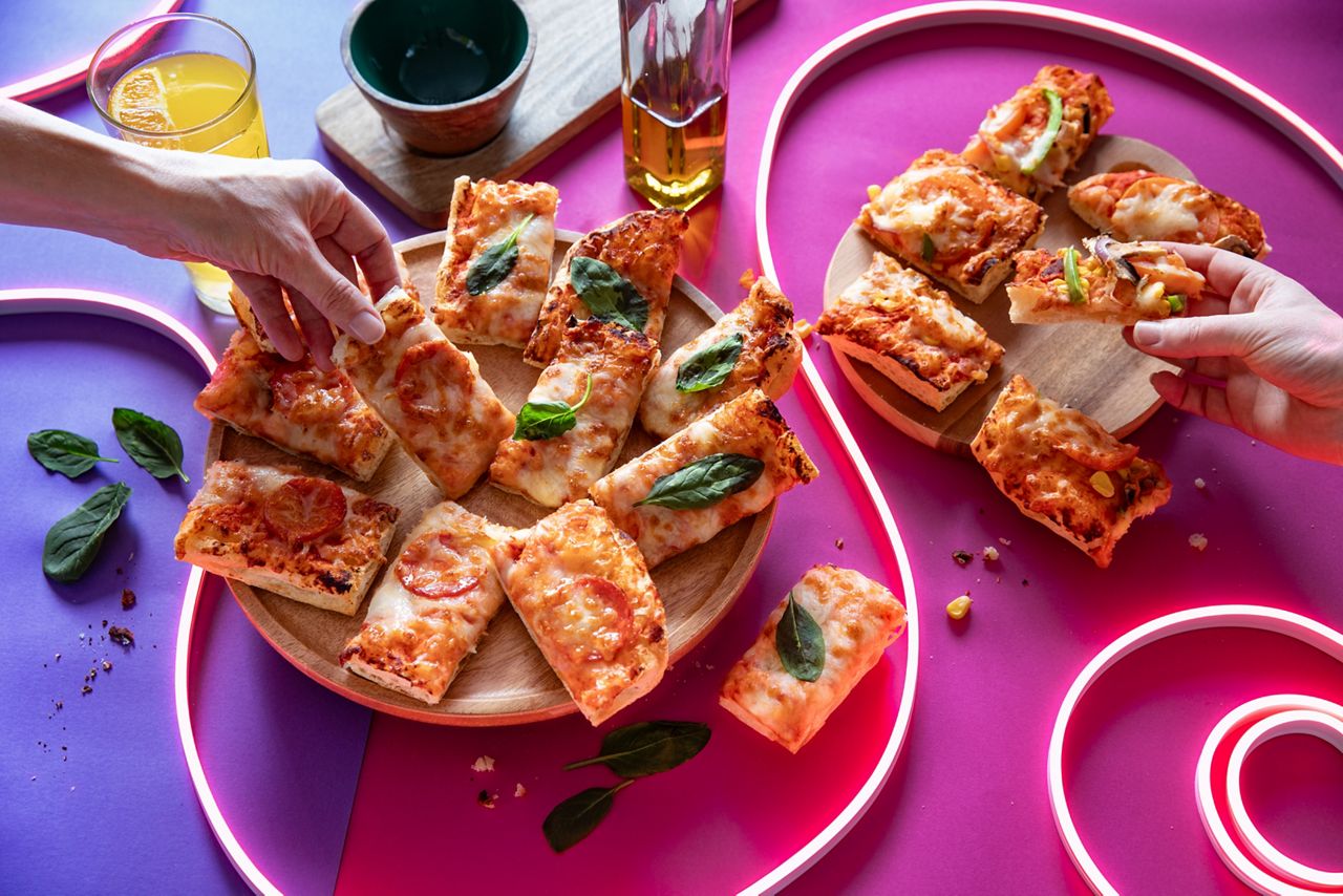 There is rectangle slices of pepperoni and margherita pizza on a wooden plate, with a soft drink in a glass next to it and a bottle of oil in the background. They all sit on a brightly coloured background of pink and purple and led light strips. There are two different hands picking up slices of pizza.