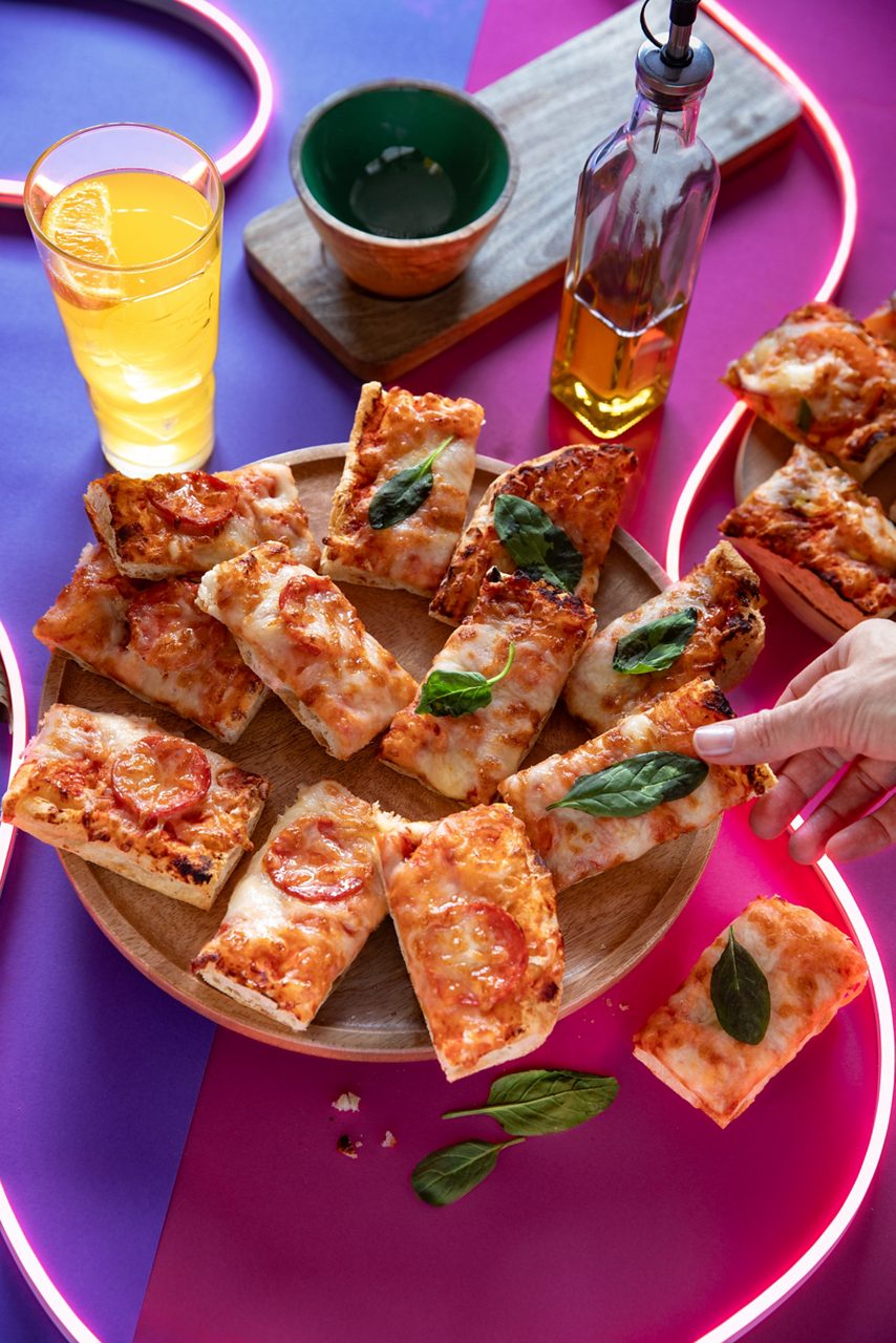 There is rectangle slices of pepperoni and margherita pizza on a wooden plate, with a soft drink in a glass next to it and a bottle of oil in the background. They all sit on a brightly coloured background of pink and purple and led light strips