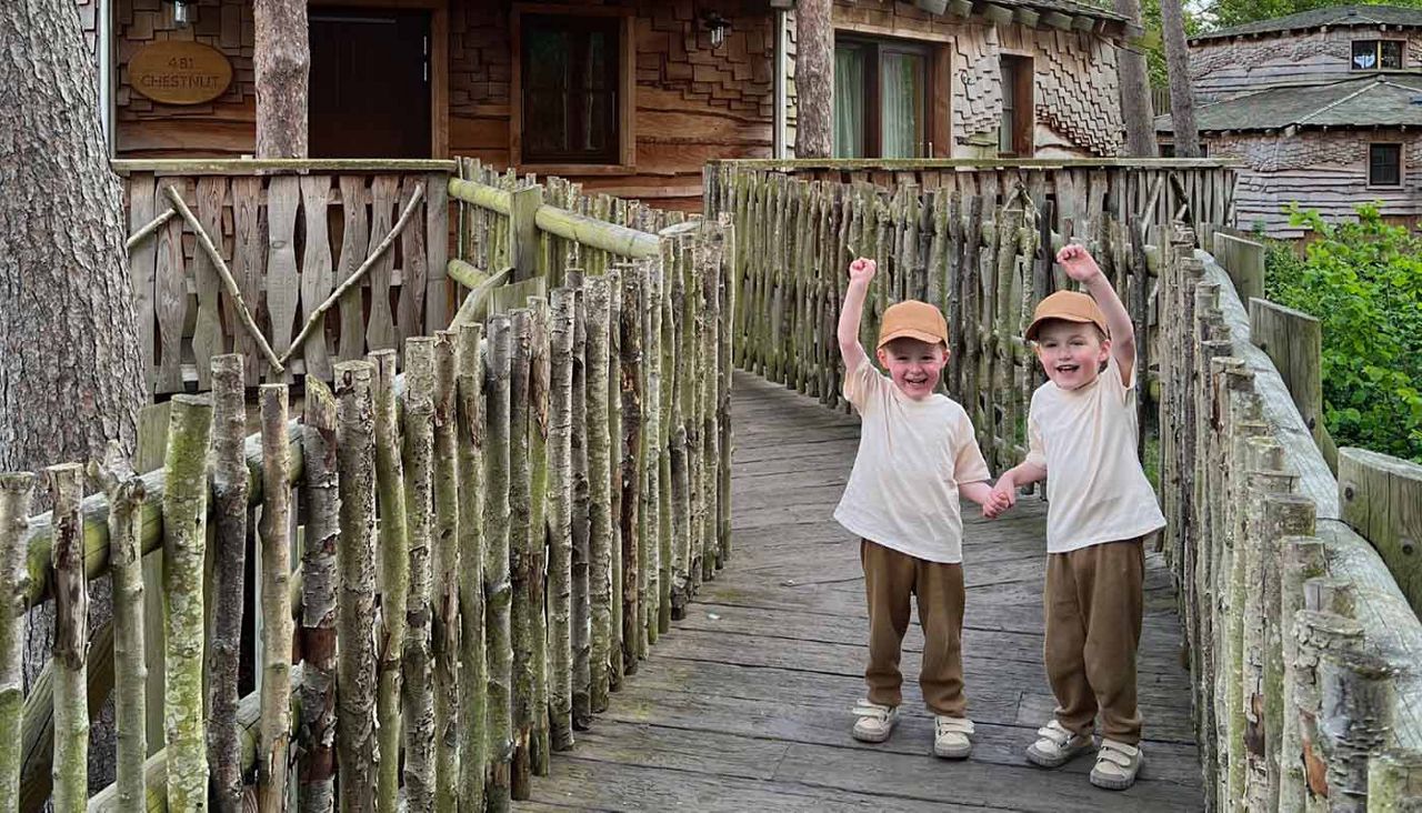 Two children hold hands and cheer, arms raised, while standing on a rustic wooden footbridge leading to treehouse cabins in a forest. Text: “CHESTNUT” and an unreadable number.