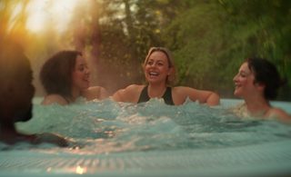 Adults laugh and chat while soaking in a bubbling hot tub, arms resting on the rim; sunlit steam rises around them in a lush, green outdoor setting.