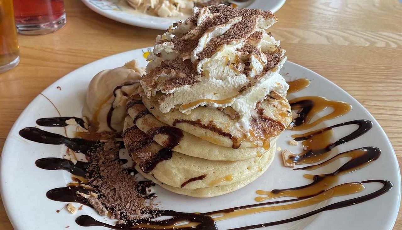 Stacked pancakes topped with whipped cream and cocoa crumble, drizzled with chocolate and caramel; a scoop of ice cream melts beside them on a white plate on a wooden table.