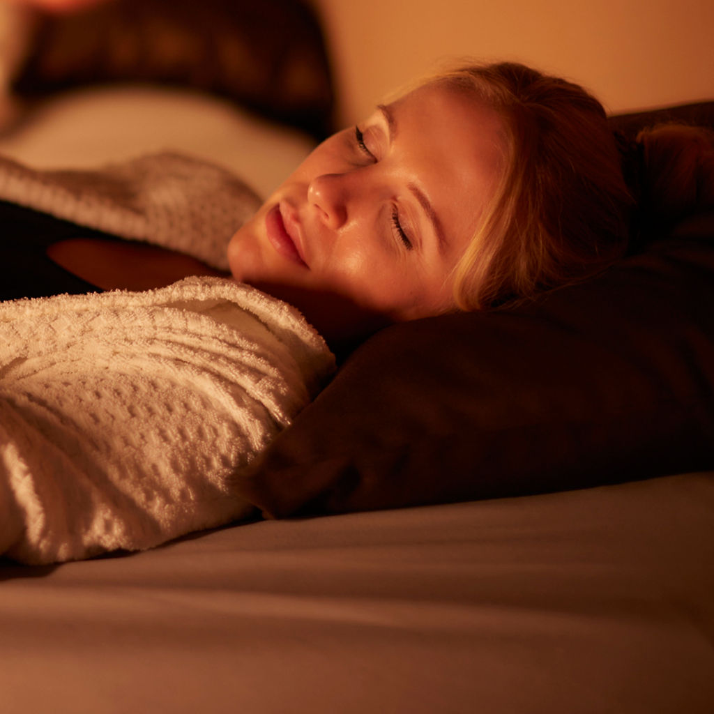 Woman sleeps on a cushioned bed, hands clasping a dark blanket, wearing a white robe; another person rests blurred in background within a warm, dimly lit spa-like relaxation room.