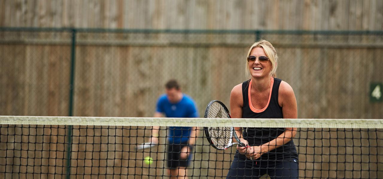Smiling woman with a tennis racket waits at the net, ready to volley; behind her, a partner in blue prepares to hit. Court with fence and a sign reading "4".