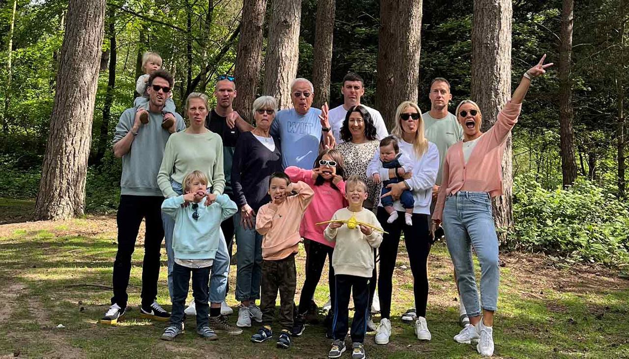 Large multigenerational group poses, some kids making silly faces, adults holding toddlers, one person on shoulders, another raises a peace sign; all standing in a sunlit forest clearing among tall trees. T-shirt text: WIT.