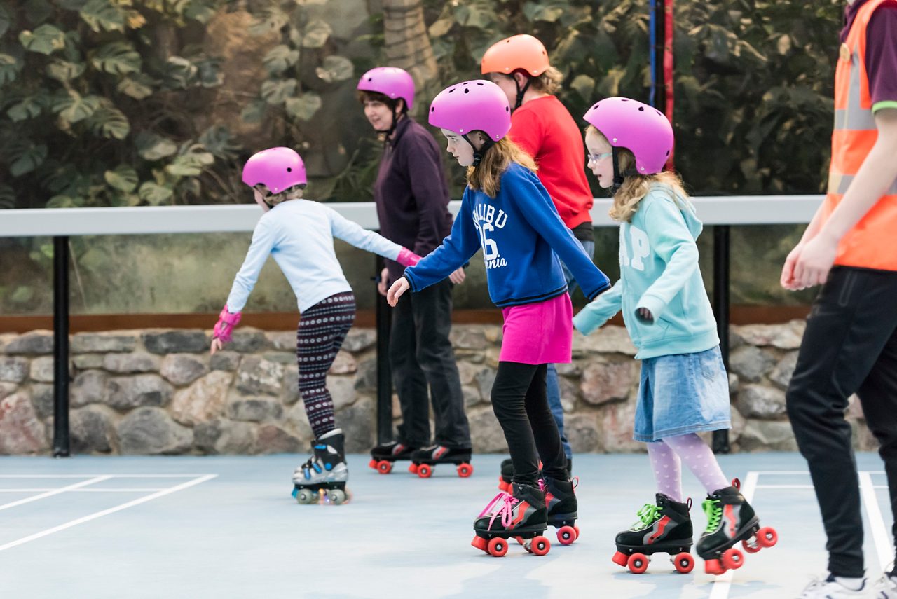 Children roller-skate, gliding in a line, wearing helmets. Indoors near a stone wall and railing. Visible text: "MALIBU 76" on a sweatshirt and "GAP" on a hoodie.