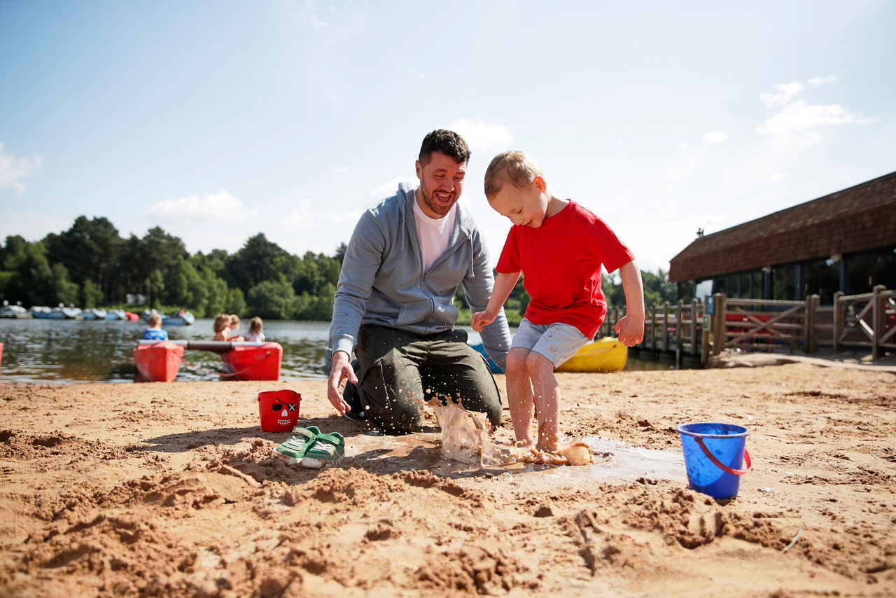 Family of four sit and eat ice lollies, chatting and smiling, on a wooden picnic bench beside a calm lake; trees, distant boats, and summer light create a relaxed setting.