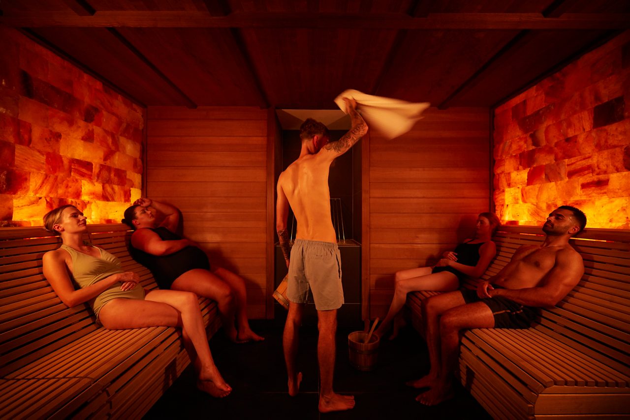 A man stood in a sauna room and four people sat on benches
