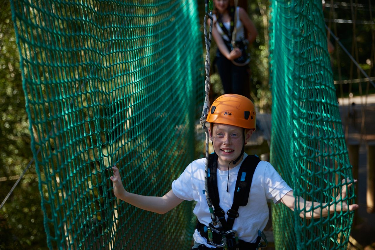 Child in orange helmet labeled “Petzl” walks across a suspended rope bridge, gripping green net sides, clipped to a harness; another participant follows behind amid trees in a treetop adventure course.