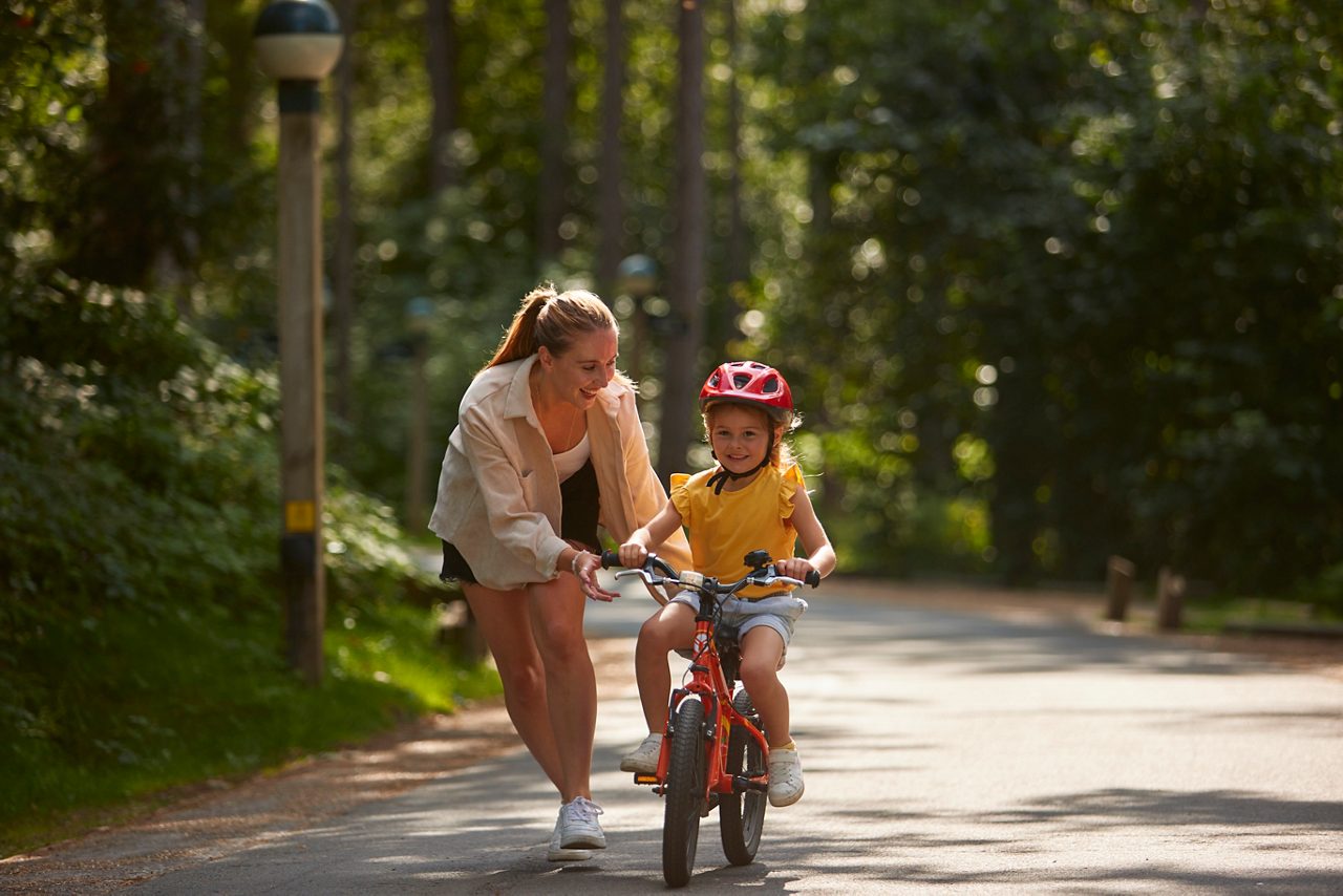 A mother helping her daughter ride a cycle through the forest.