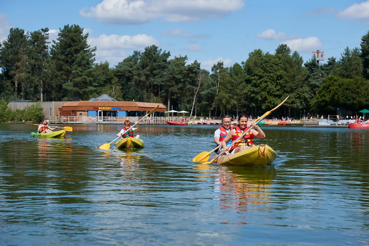 Kayakers paddle yellow boats, wearing life jackets, across a calm lake; behind them a wooded shoreline and a waterside building labeled THE PANCAKE HOUSE under a blue, partly cloudy sky.