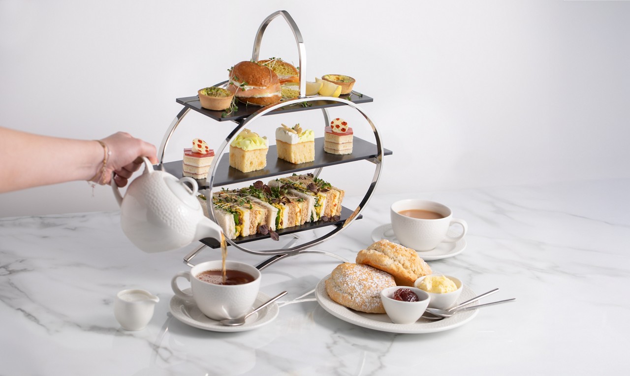 image of a hand pouring a pot of tea into a cup, infront of a afternoon tea stand with cakes and sandwiches on 