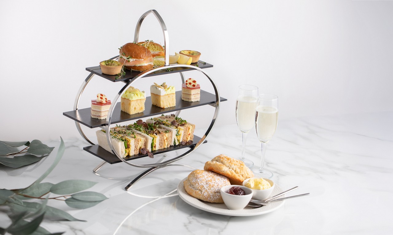 afternoon tea stand with cakes and sandwiches next to a plate with scones and two glasses of prosecco