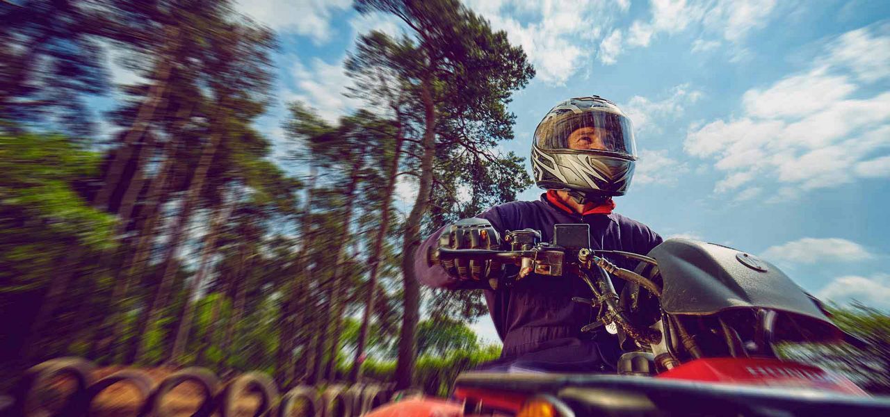 Helmeted rider controls a red ATV, accelerating along a wooded trail; motion blur streaks tall pines and blue sky, with stacked tires edging the path.