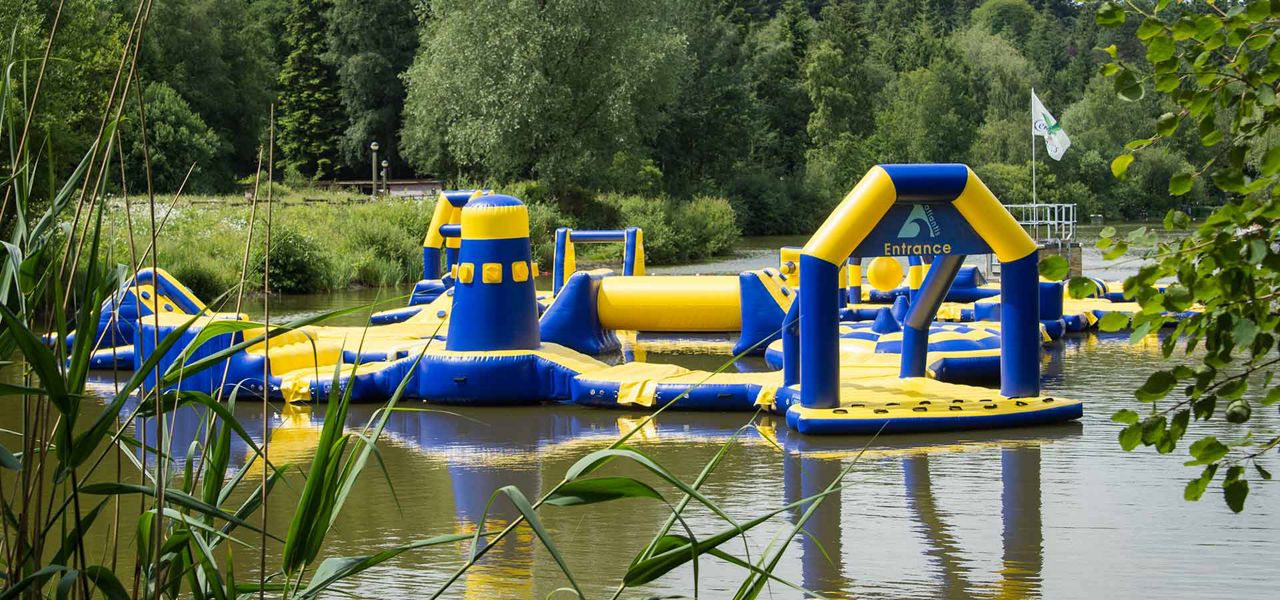 Inflatable water obstacle course floats, inviting entry through an arch labeled "Entrance", within a calm lake; yellow-and-blue towers, slides, and paths sit amid reeds and surrounding leafy trees.
