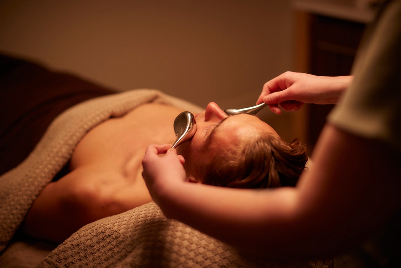 Client receiving treatment as therapist glides two metal spoons over closed eyes and forehead, on a massage table under a textured beige blanket in warm, dim spa lighting.
