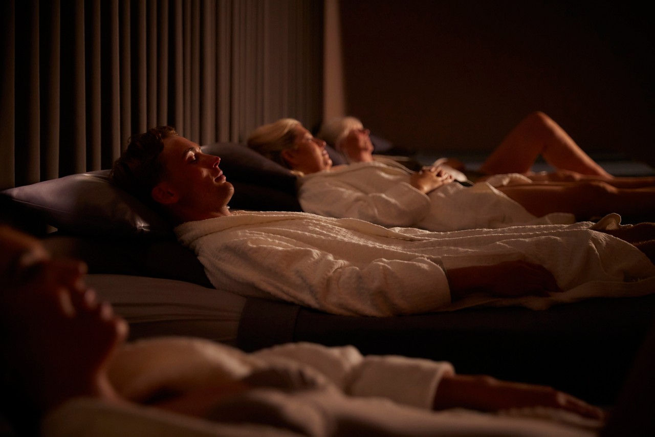 people in spa robes lying on padded daybeds resting. Warm, low lighting and ribbed wall panels create a calm, dim relaxation-room ambiance.