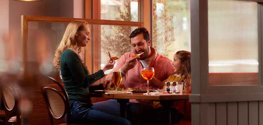 Family eats pizza at a booth in a cozy restaurant. Adults hold beer and an orange cocktail while a child smiles beside them amid warm lighting and wooden partitions.