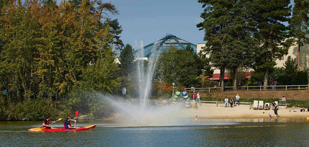 Kayakers paddle a red kayak across a lake, passing a tall water fountain; onshore, people relax on a sandy beach near trees, stacked boats, and a building with CAFE ROUGE awnings.