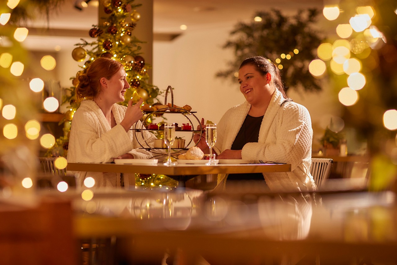 two women wearing white plush spa robes smiling at a table while enjoying a festive afternoon tea with champagne near a lit Christmas tree