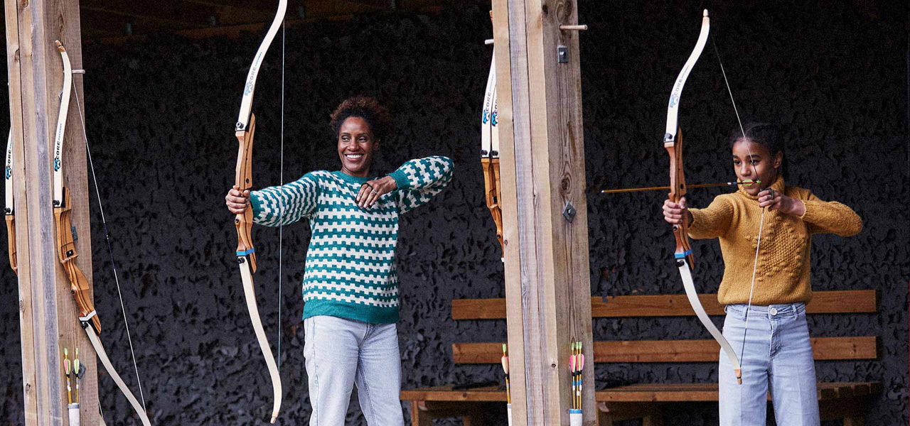 Archers shoot; one draws an arrow while another holds a bow smiling; covered wooden range with hanging bows, benches, and quivers. Text: CORE on multiple bows.