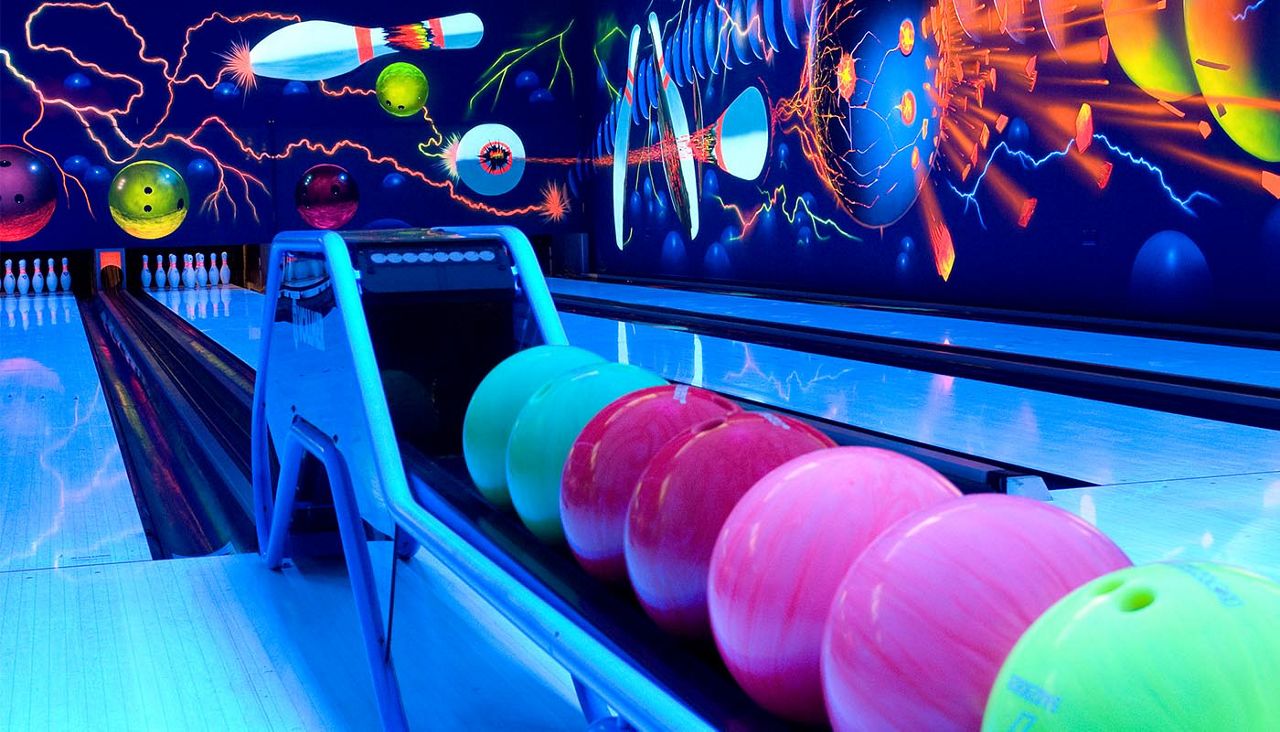 Neon bowling balls rest on a ball return, awaiting play, in a blacklight bowling alley; glowing lanes lead to pins, with vivid cosmic wall art of pins, balls, and lightning.