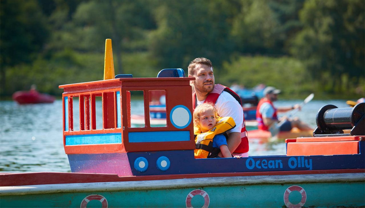 Adult and child wearing life jackets sit in a colorful toy-like boat, steering and looking ahead, on a calm lake with other boaters nearby; boat name text: Ocean Olly.