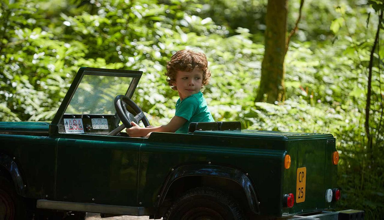 Child drives a small green toy jeep, turning the steering wheel and looking back; surrounding sunlit forest path with lush greenery. License plate reads CP 25.