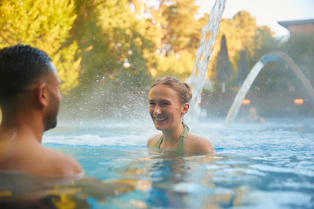two people in an outdoor pool if water falling behind them and forest trees in the background