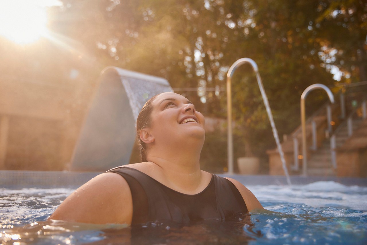 A lady in the outdoor pool in front of a water fountain looking at the sky with the sun in the background.