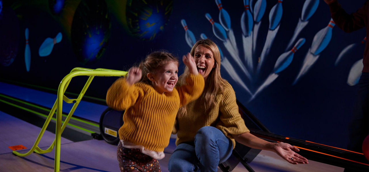 Child jumps with raised arms, celebrating. Woman kneels beside, smiling. Context: dimly lit bowling alley with neon lanes, pins mural, and a bright green bowling ramp for assistance.