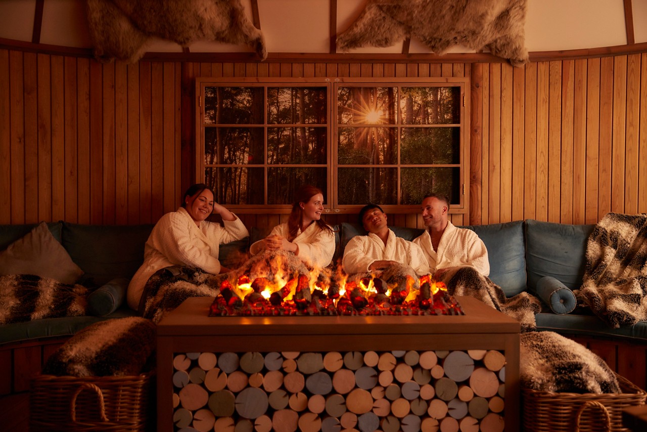 a group of people in spa robes sat on a sofa infront of a fire